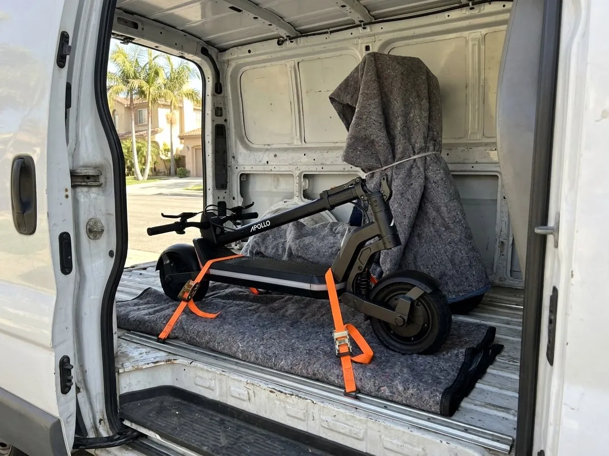 Folded electric scooter strapped down on moving blankets inside a cargo van on a sunny residential street